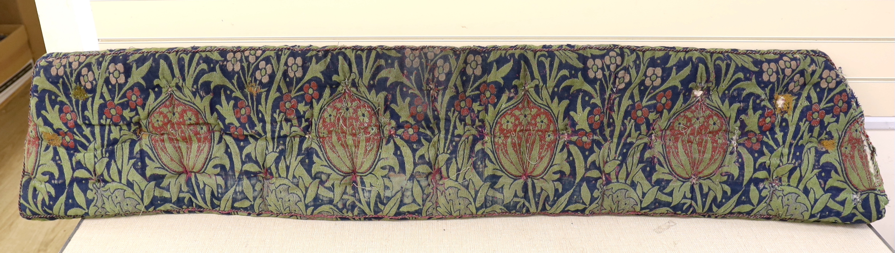 A William Morris Elmore, wool and mohair woven panel by J H Dearle circa 1900, used to cover a buttoned and braided window seat cushion, 196cm wide (at widest point) x 38cm deep, Please note this lot attracts an addition